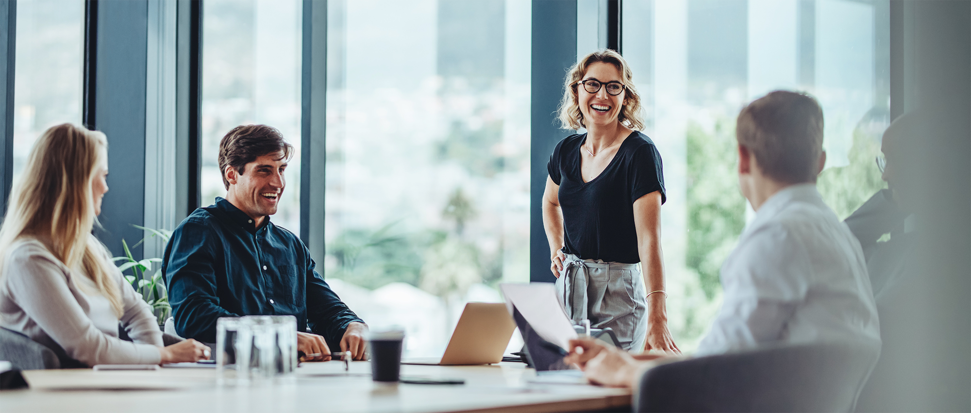 A group of four business professionals is in a meeting room with large windows overlooking a city. A woman with glasses and a black shirt stands smiling at the center. To her left, a man in a dark blue shirt is laughing, and a woman is looking at him. To her right, a person in a white shirt is turned towards her. A laptop and coffee cup are on the table.