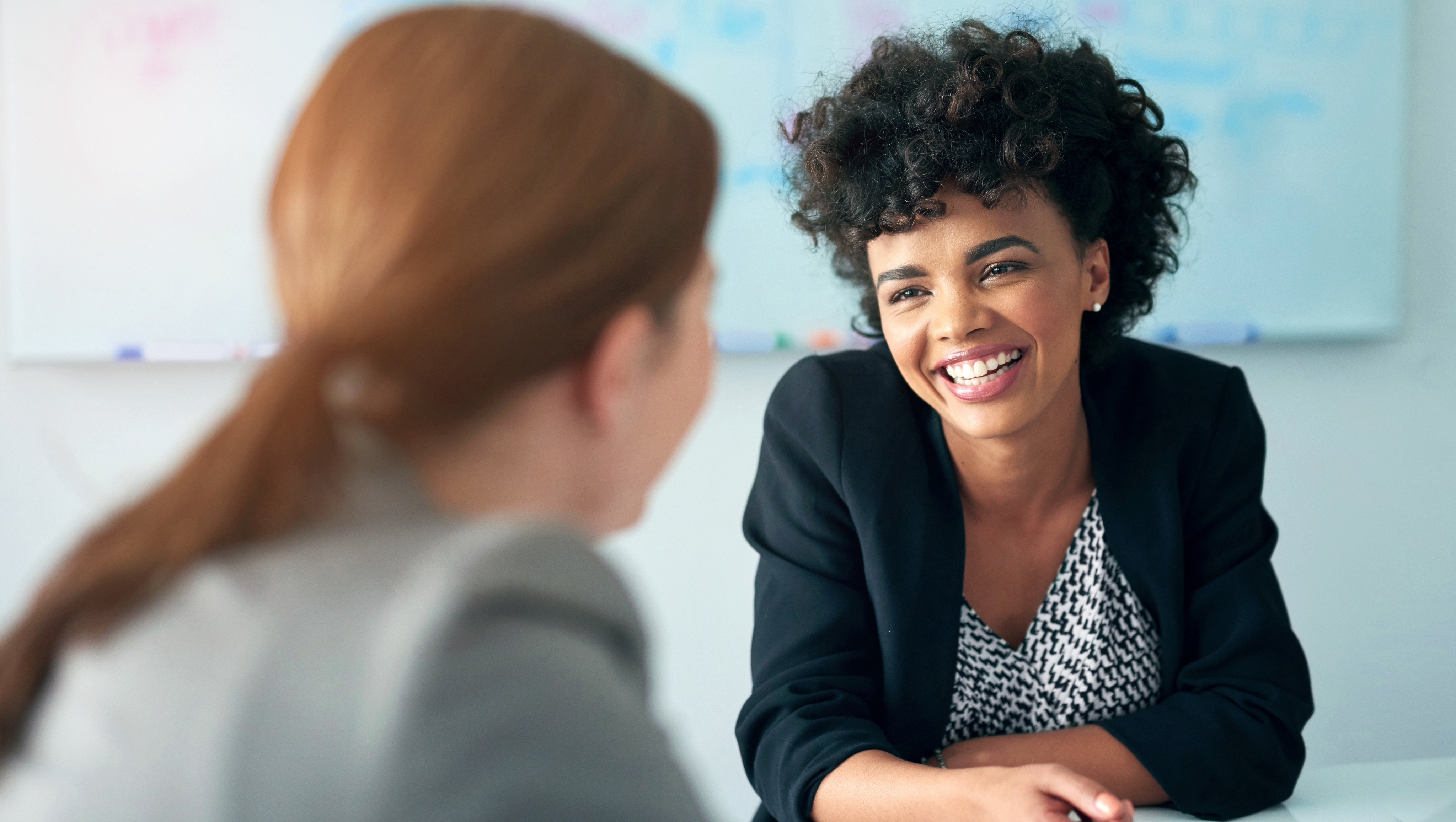 Two women are in a professional setting, possibly an office or a meeting room. One woman is in focus, smiling warmly and looking at the other woman, who is partially visible with her back to the camera. The woman in focus has a dark, curly hairstyle and is wearing a black blazer over a patterned top. The other woman, out of focus, has light-colored hair pulled back in a ponytail and is wearing a gray blazer. A whiteboard with some text and a colorful chart is visible in the blurry background.