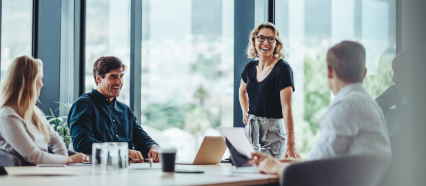 A group of four business professionals is in a meeting room with large windows overlooking a city. A woman with glasses and a black shirt stands smiling at the center. To her left, a man in a dark blue shirt is laughing, and a woman is looking at him. To her right, a person in a white shirt is turned towards her. A laptop and coffee cup are on the table.